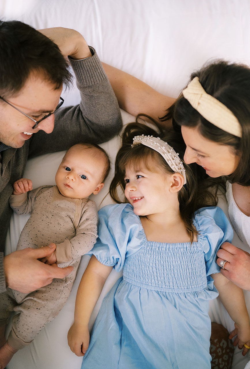 A family of four consisting of young parents, their toddler and baby, poses for a photo on a bed